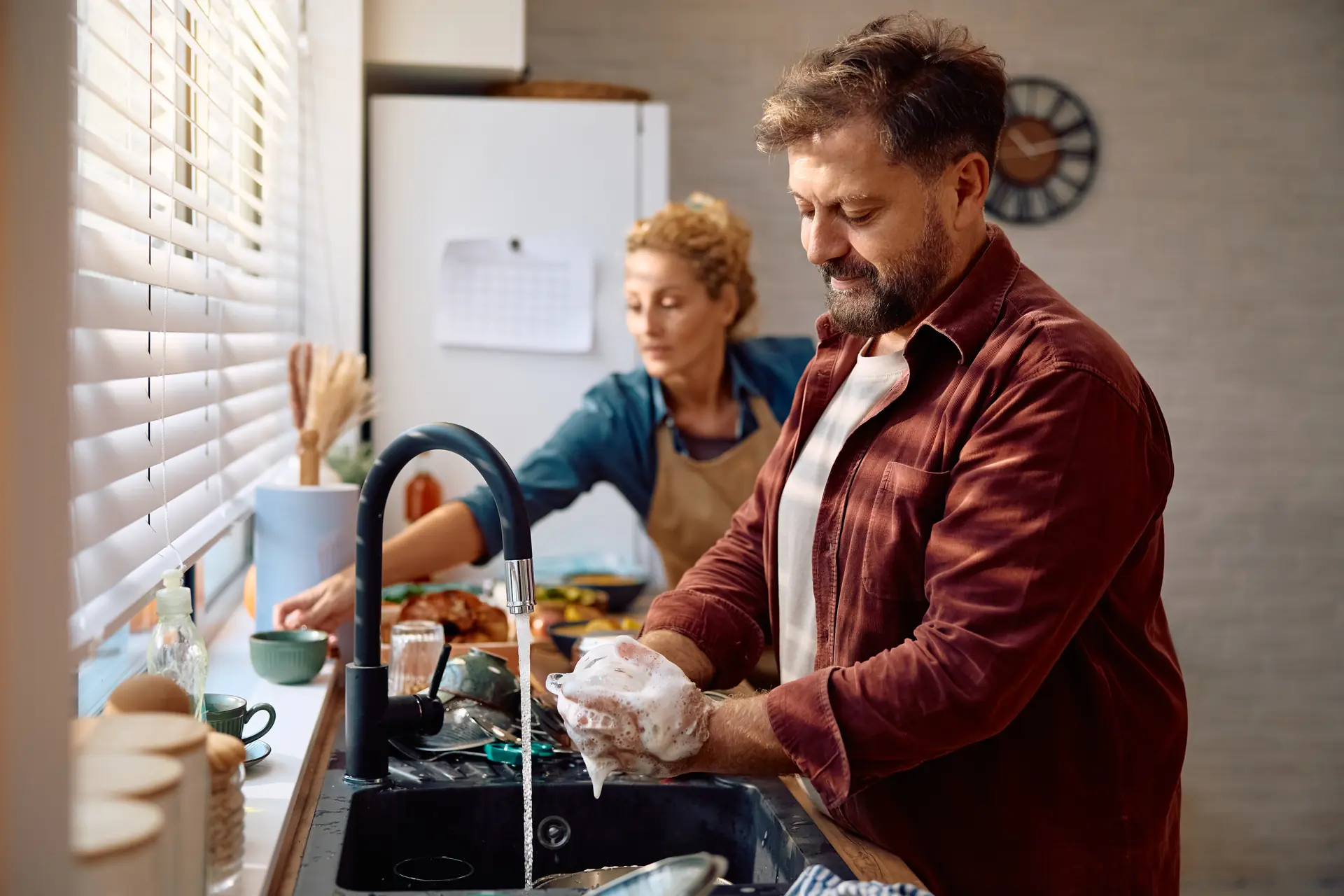 Man washing dishes at kitchen sink while woman prepares food in background
