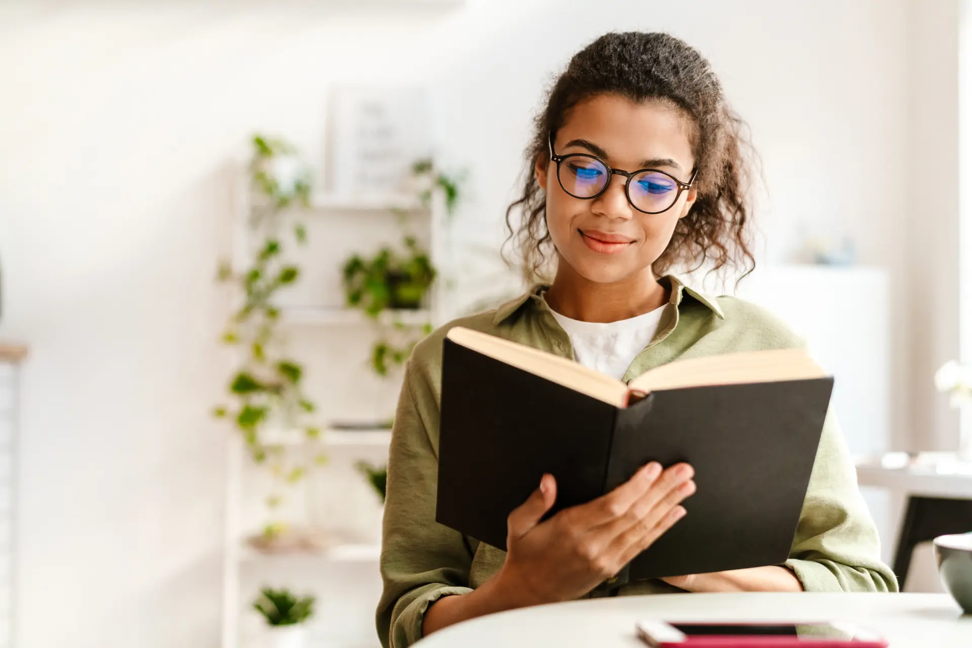 Woman with glasses reading a book indoors with plants in background