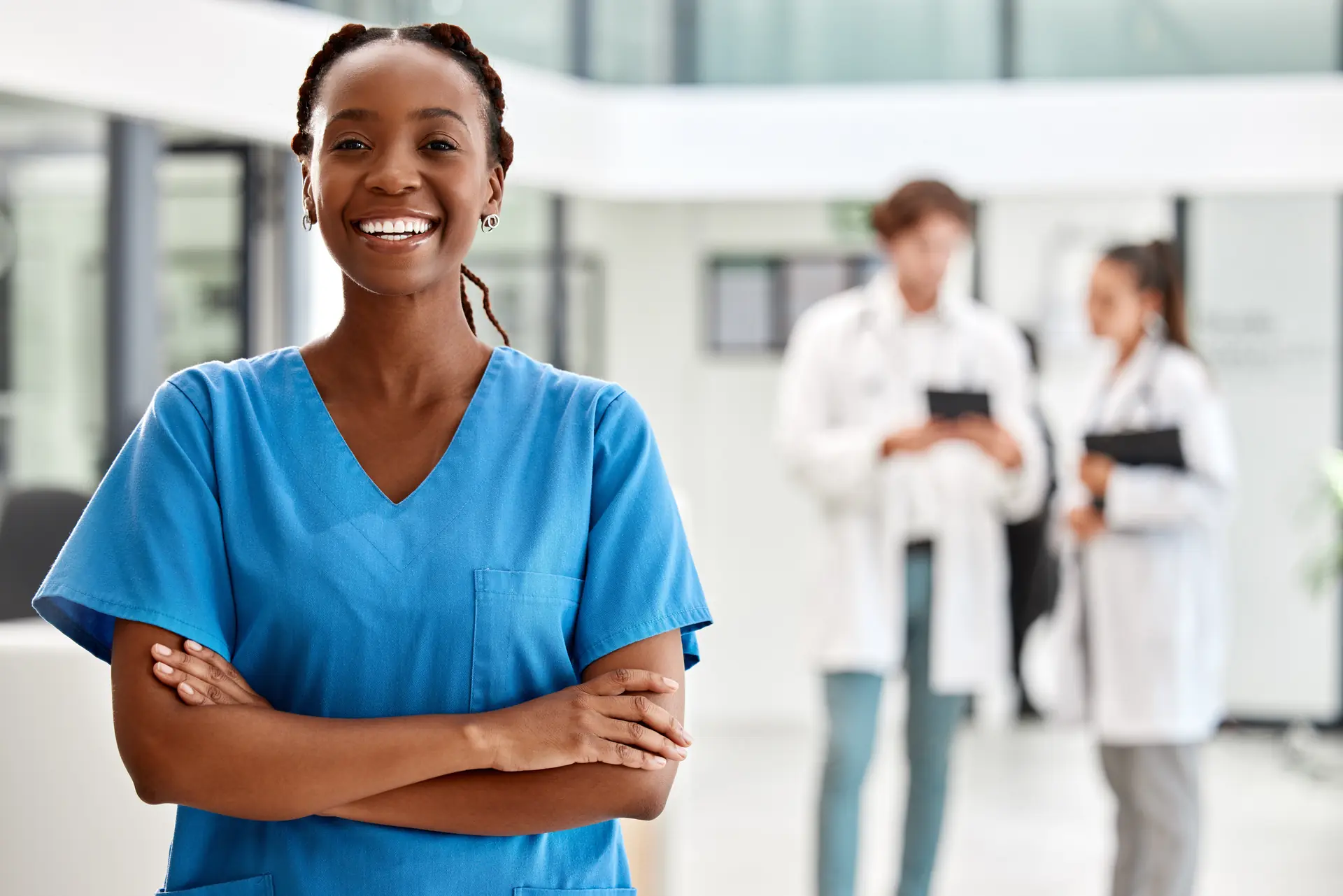 Smiling nurse in blue scrubs with arms crossed, doctors talking in background