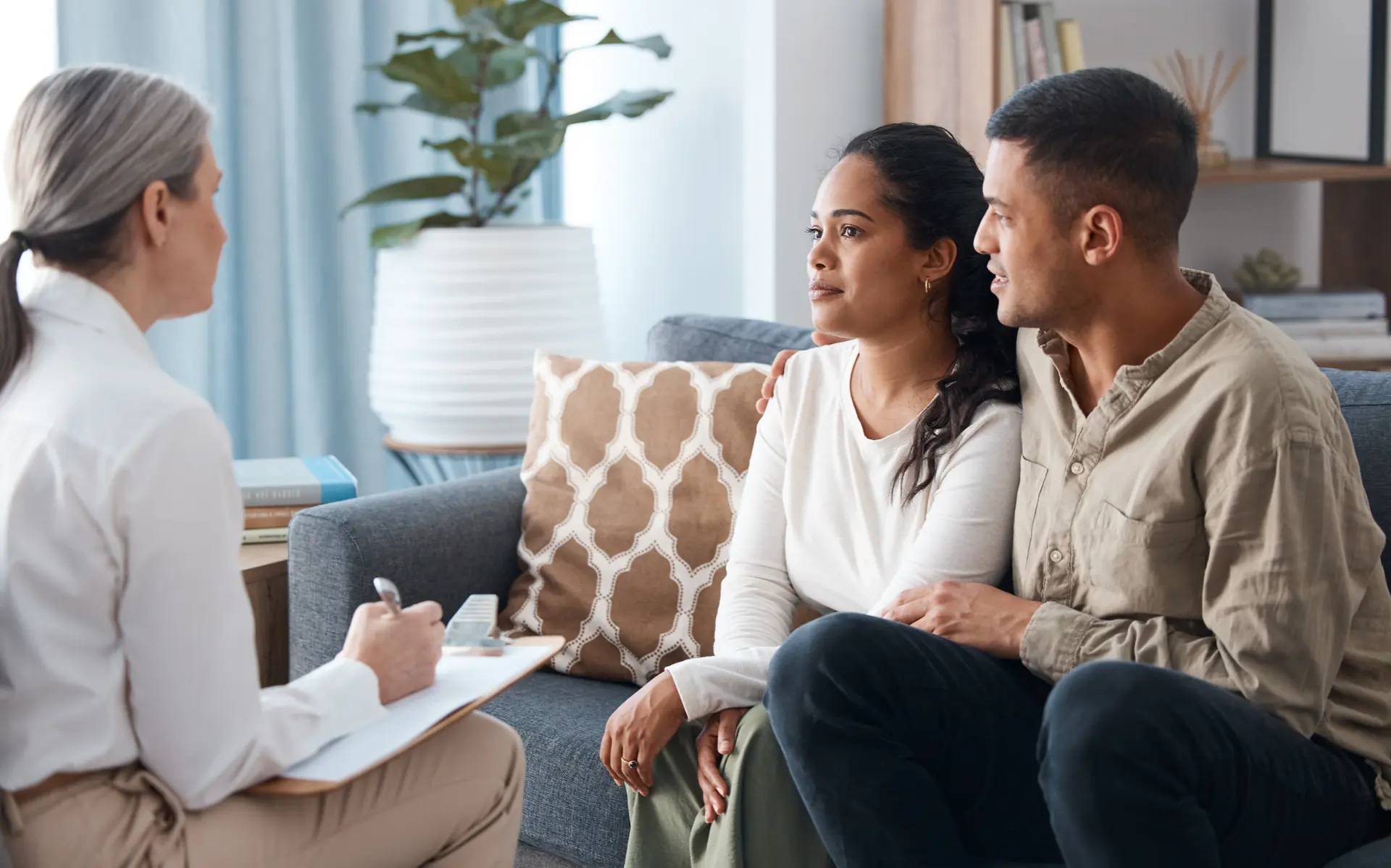 Couple sitting on couch talking with therapist holding clipboard in a bright office.