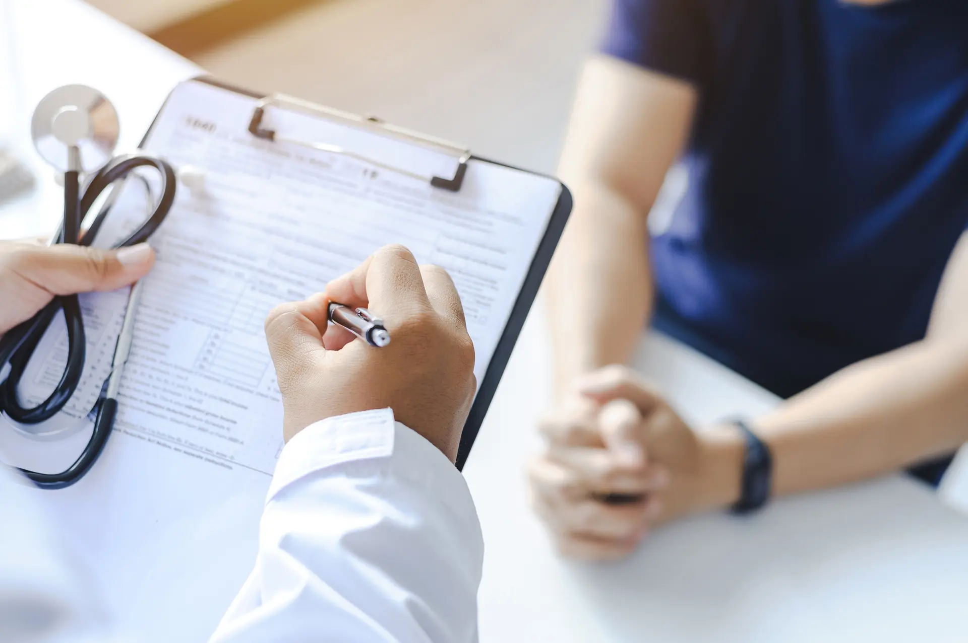 Doctor writing notes on clipboard while patient sits with hands clasped nearby